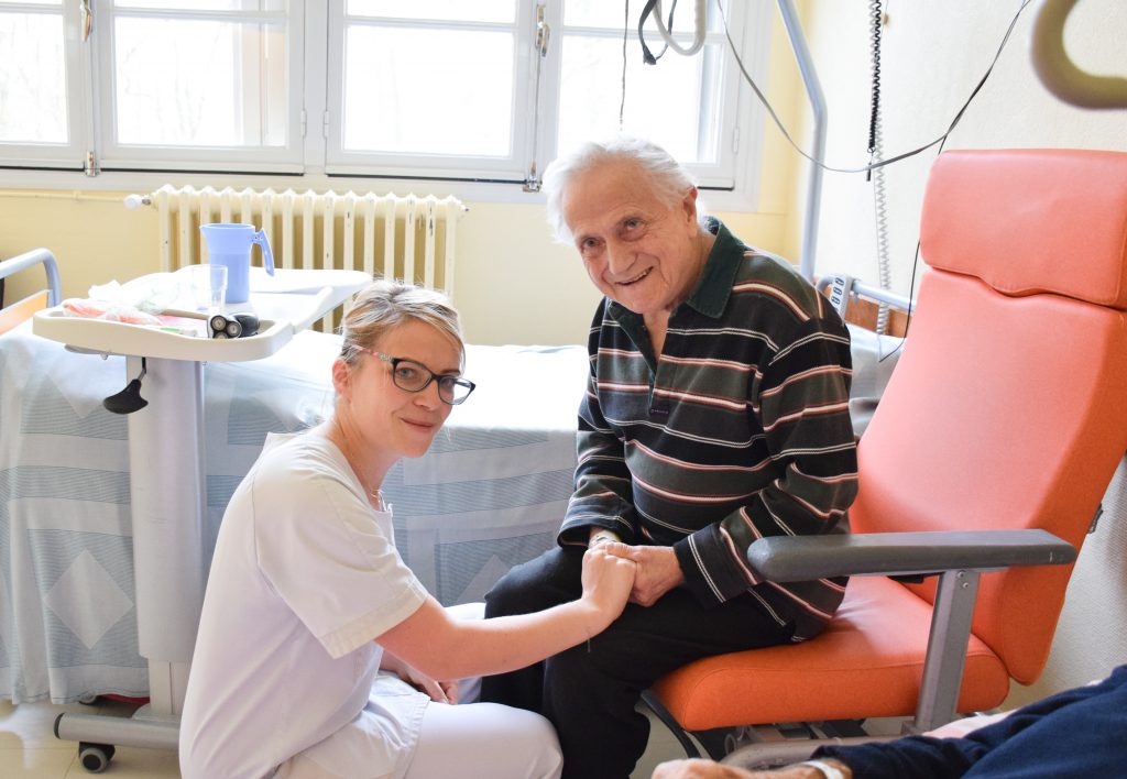 Une infirmière est accroupie et tient la main d'un patient installé sur le fauteuil de sa chambre.